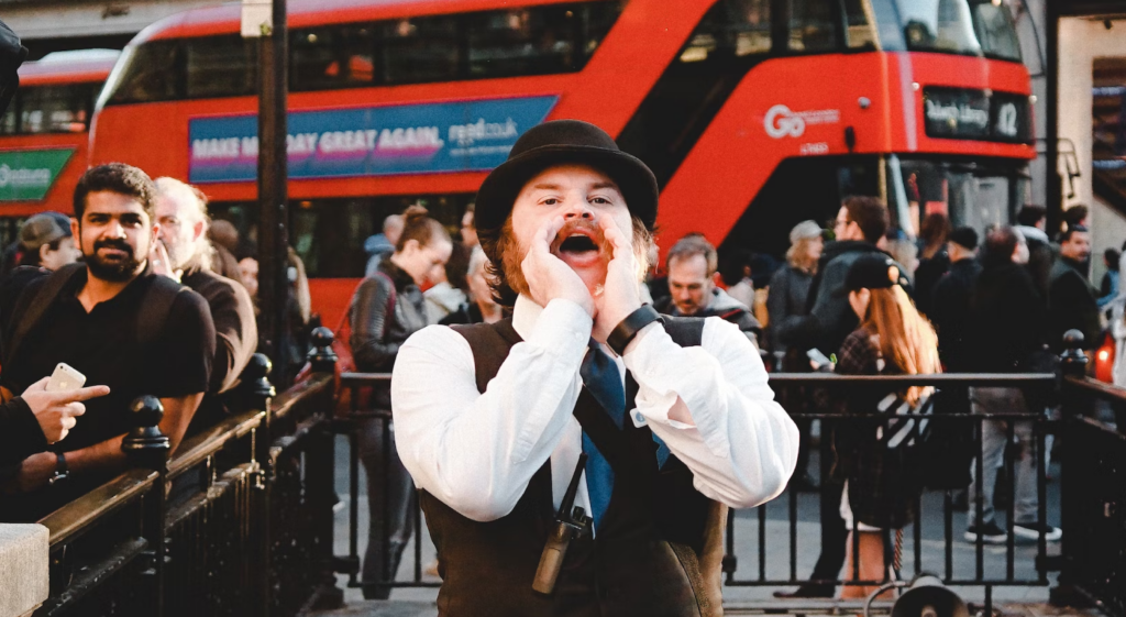 image showing man dressed in Victorian-esqu attire shouting in front of a red London bus